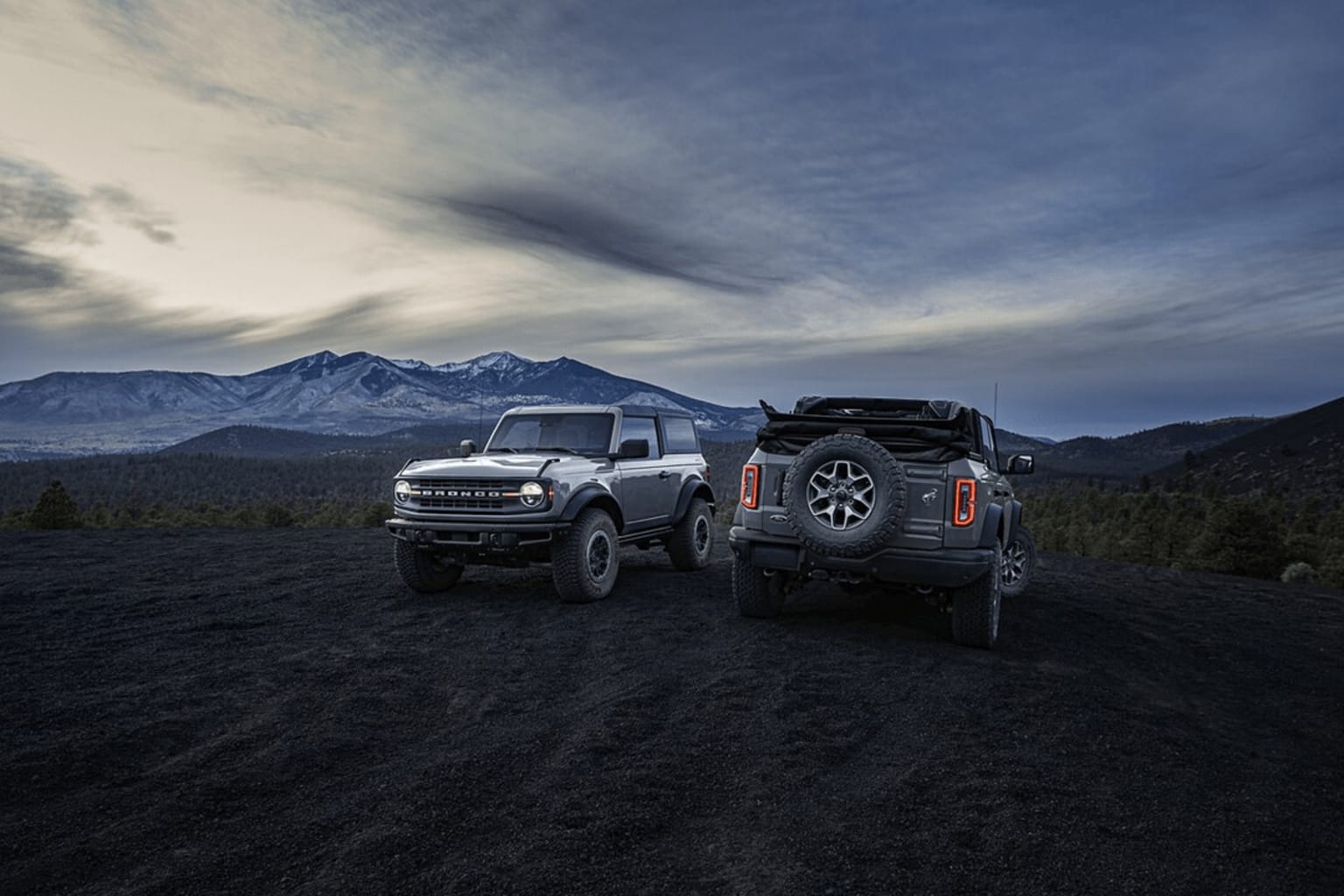The image shows an off-road vehicle in a mountainous landscape with snow-capped peaks in the background and a dramatic cloudy sky overhead.
