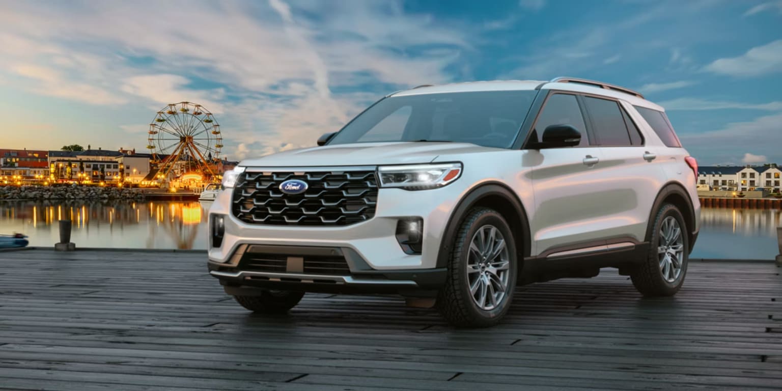 White SUV with a distinctive grille design parked on a wooden dock, with amusement park attractions visible in the background against a colorful sunset sky.