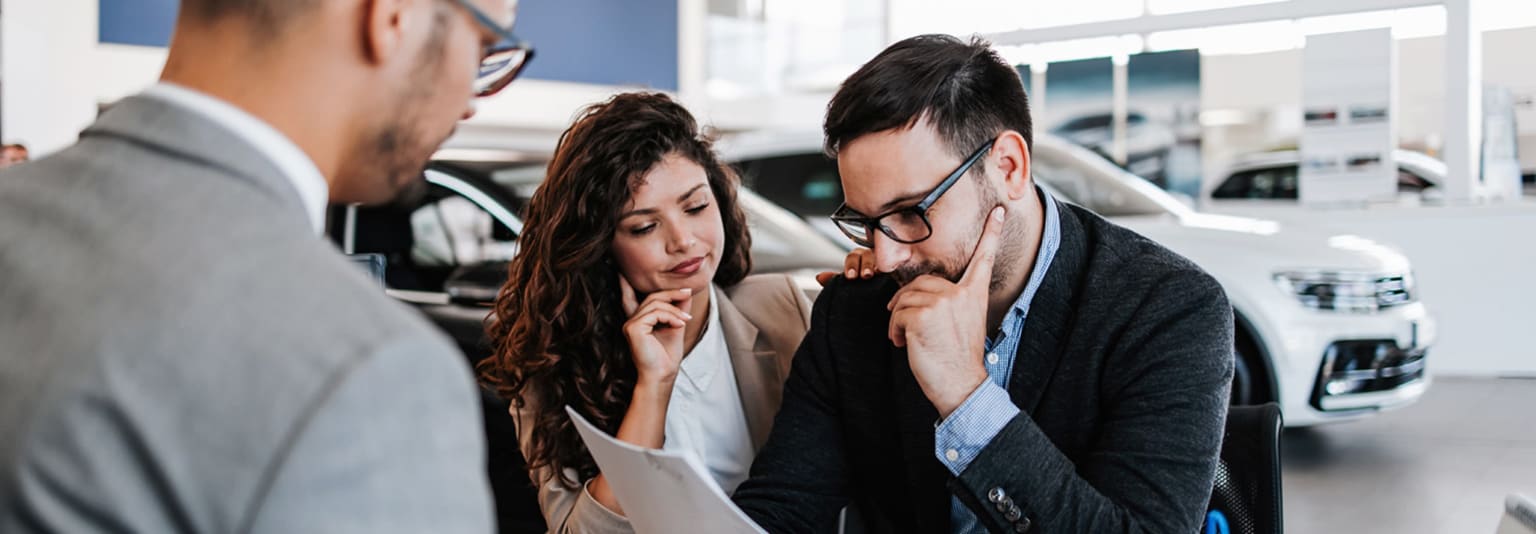Salesperson assisting customers in a car dealership showroom with new cars in the background.