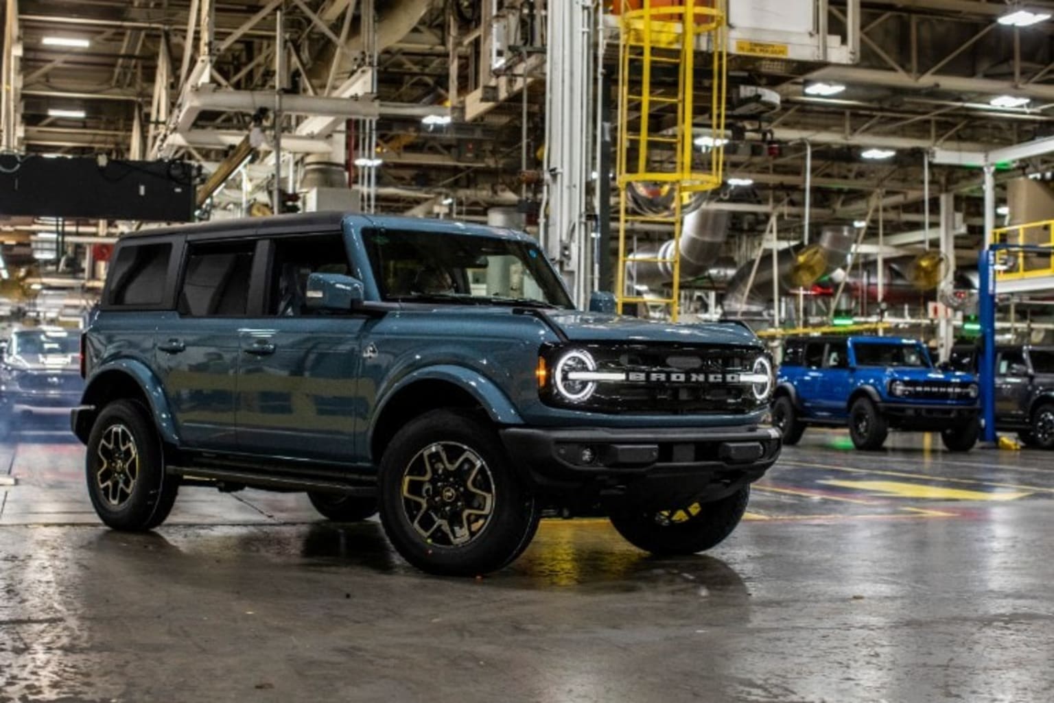 a blue ford bronco is parked in a large building with other trucks in the background and a yellow truck in the foreground