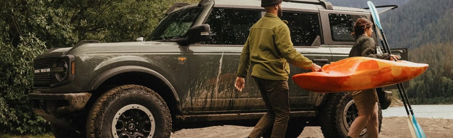 A couple carries a kayak and paddle next to a muddy SUV with a lake and mountains in the background.