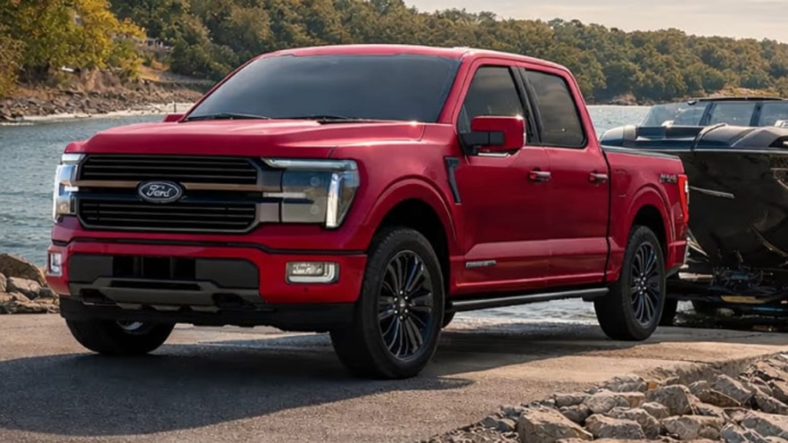 A red pickup truck is parked on a rocky shore with a boat trailer and a body of water in the background.