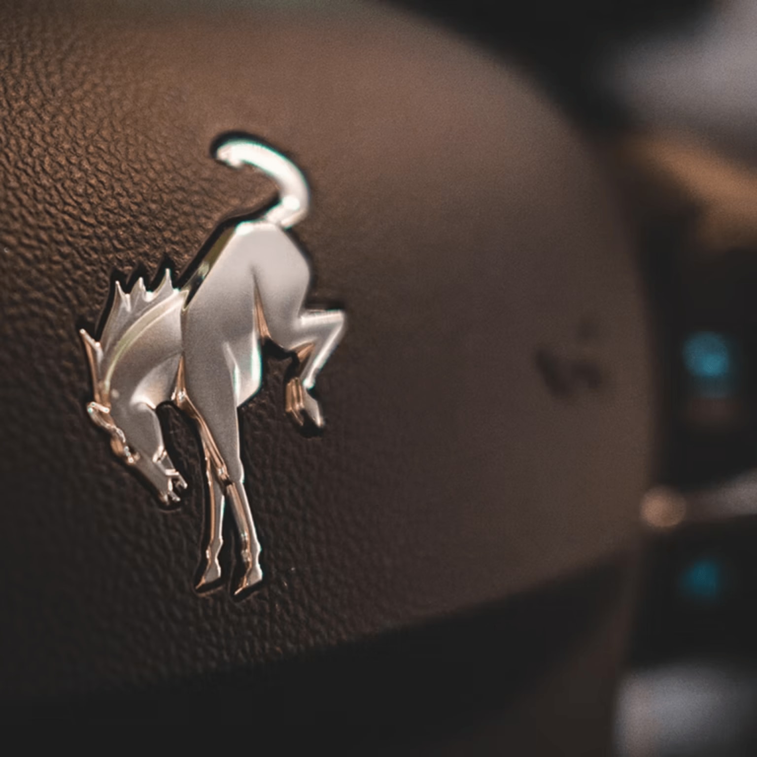 A silver bronco emblem is prominently displayed on a dark, textured surface in the foreground, with a blurred background suggesting a car interior.