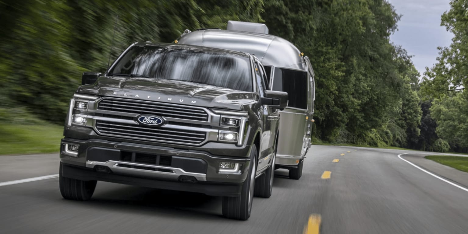 A silver-colored pickup truck driving down a winding road lined with lush, green trees.