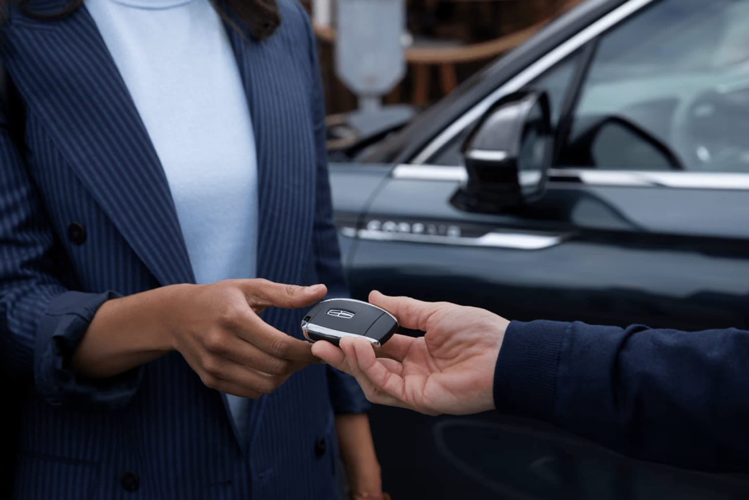 A man in a navy blue suit holding a small device, likely a car key fob, in front of a parked car in the background.