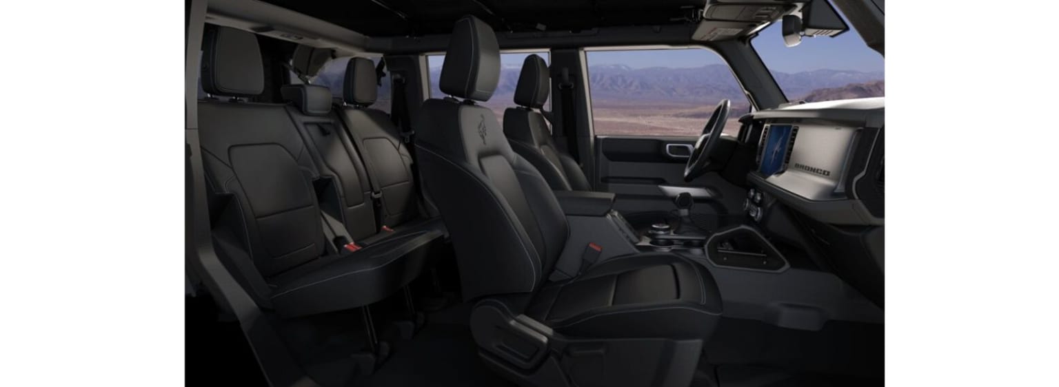 The image shows the interior of a spacious vehicle with multiple rows of seats and a large windshield providing a panoramic view of a mountainous landscape in the background.