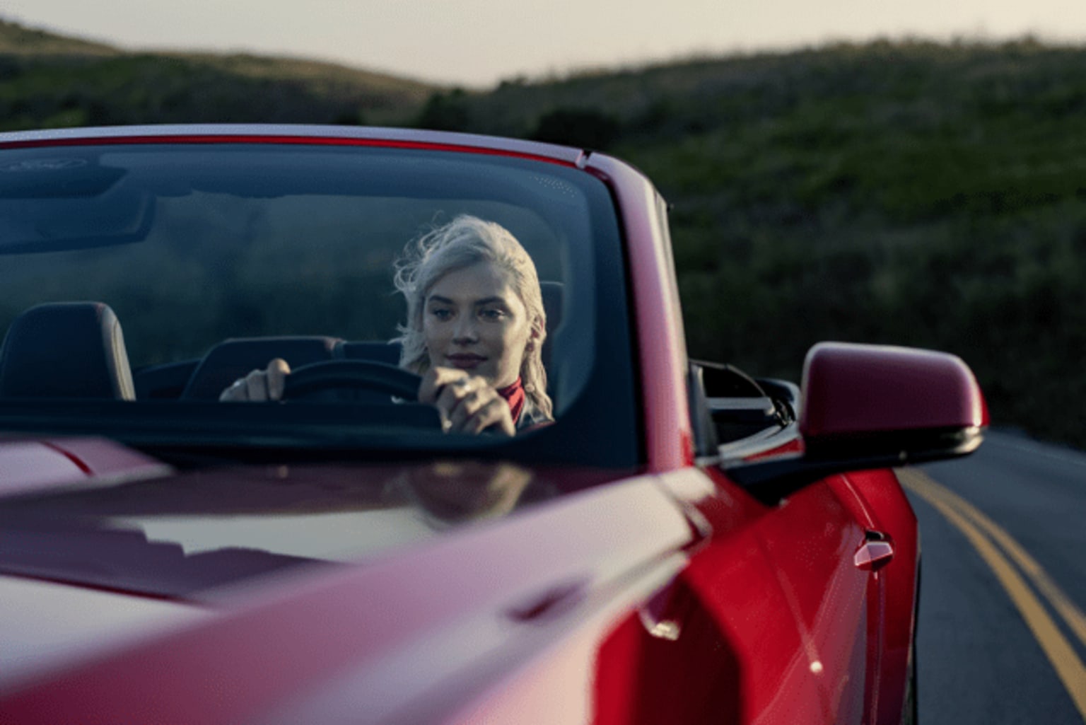 A person, likely a woman, sits in the driver's seat of a red car with a mountainous, forested landscape in the background.