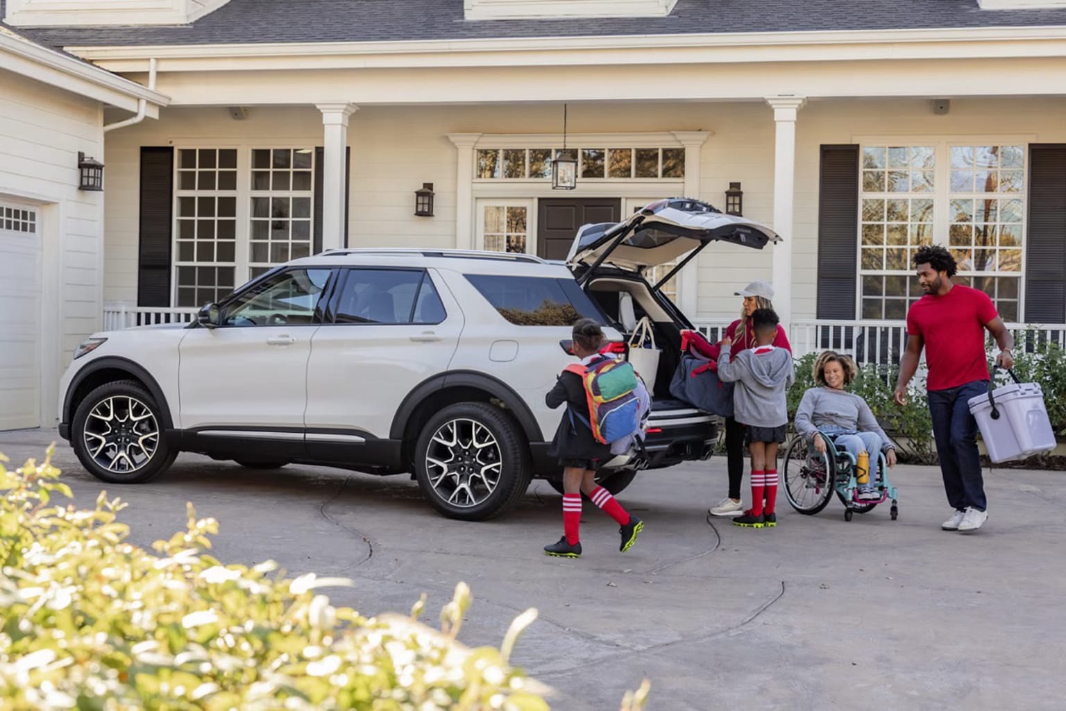 A family is gathered around a large SUV in the driveway of a suburban home, with the adults and children loading luggage and supplies into the vehicle's open trunk.