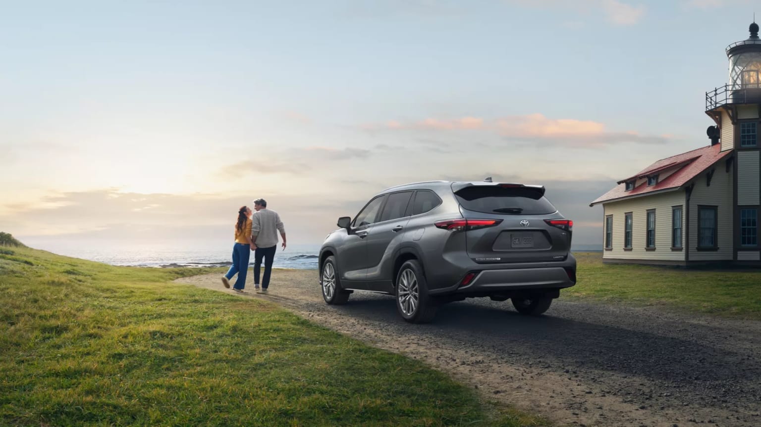 Gray SUV parked on a dirt road with a person in the foreground and a lighthouse in the background with scenic landscape.