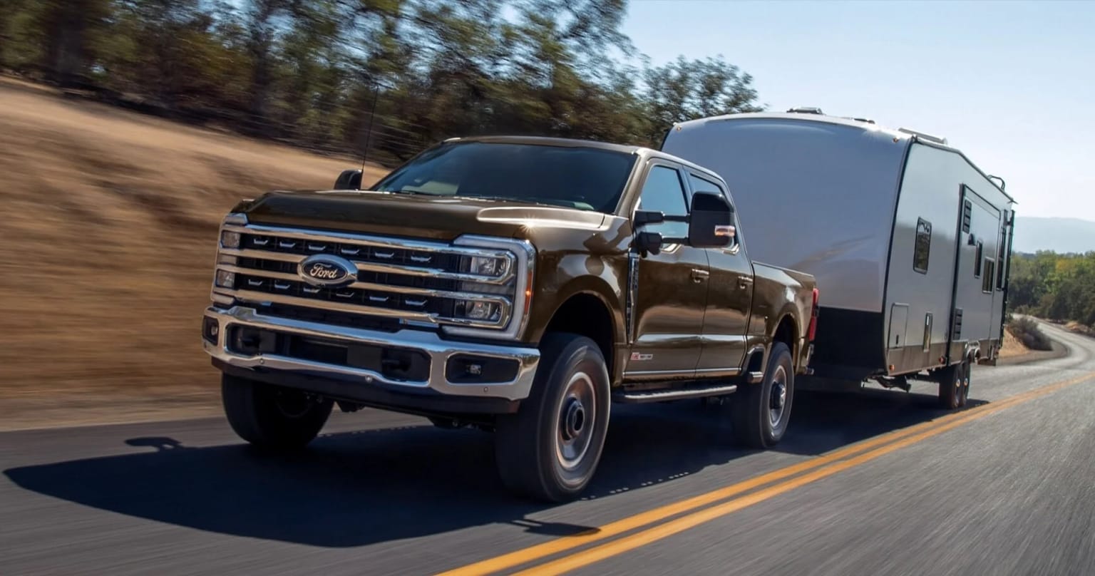 A large pickup truck towing a camper trailer is driving down a dirt road surrounded by a forested landscape.