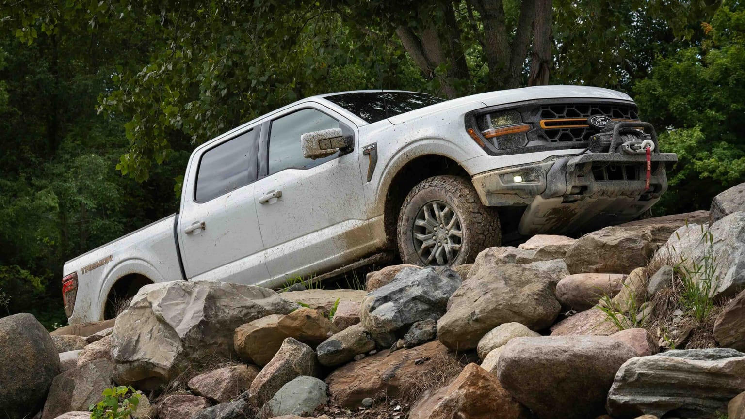A white pickup truck climbs over a rocky foreground against a backdrop of lush green trees.