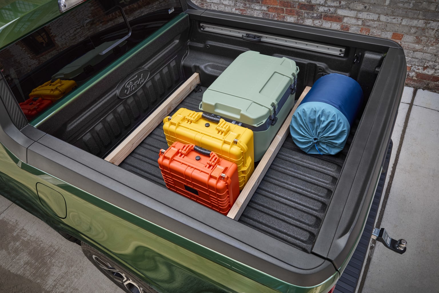 The image shows the open trunk of a vehicle filled with various colorful luggage and bags, including a green suitcase, a yellow hard-case, and a blue rolled-up item, all against a backdrop of a brick wall.