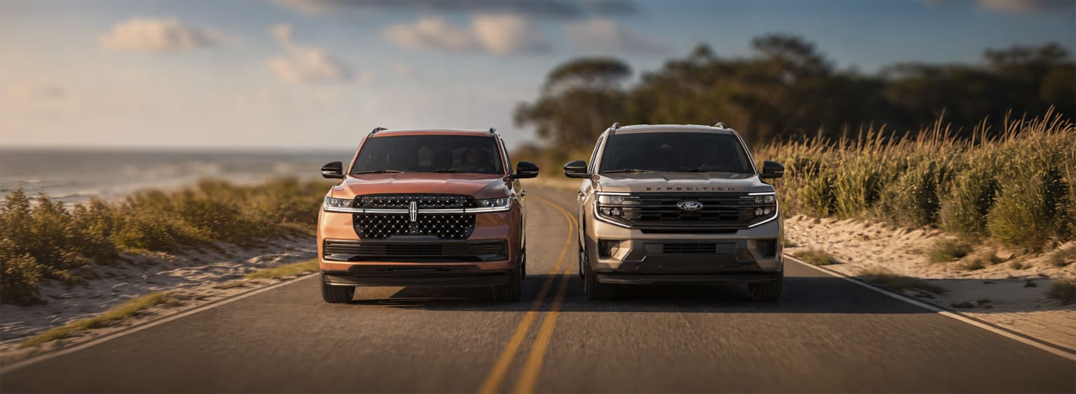 Two large SUV vehicles parked on a rural road, surrounded by lush vegetation and a scenic landscape in the background.