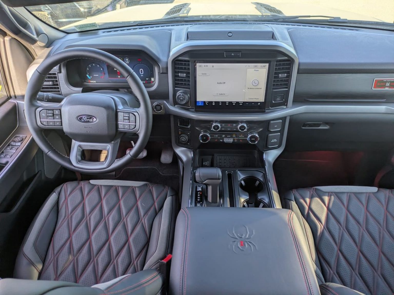 Foreground image of quilted leather seats with red stitching and a central console with a spider emblem. Background shows a modern truck dashboard with a large touchscreen and digital instrument cluster.