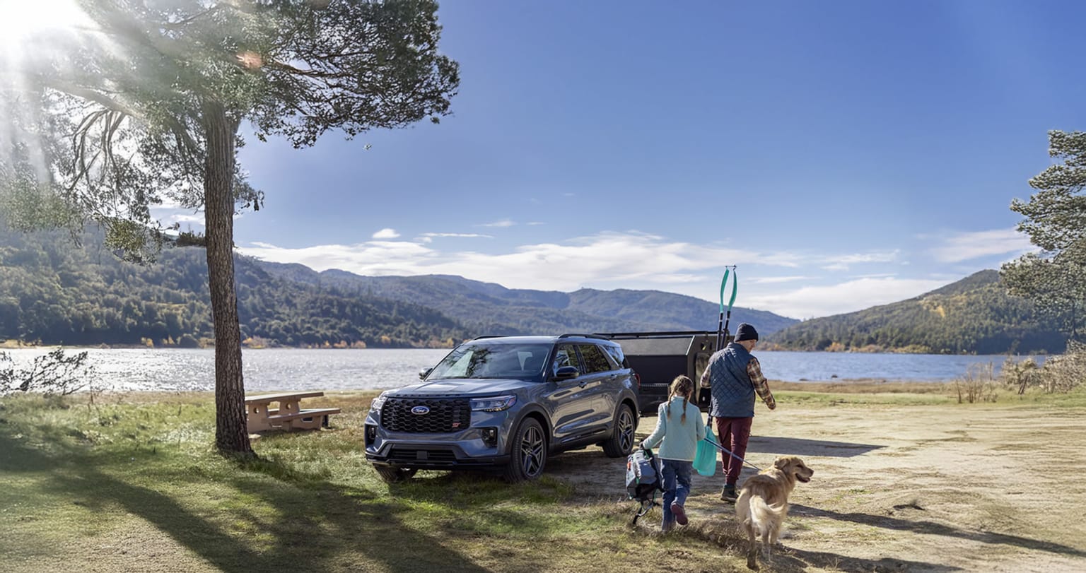 Off-road 2025 Ford Explorer parked near a lake with lush greenery and mountains in the background, person walking with dog.