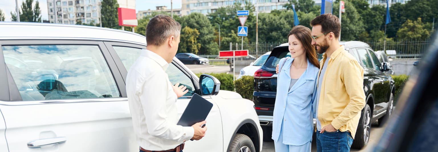 A car salesman showing a white SUV to a couple in a car lot with buildings and trees in the background.