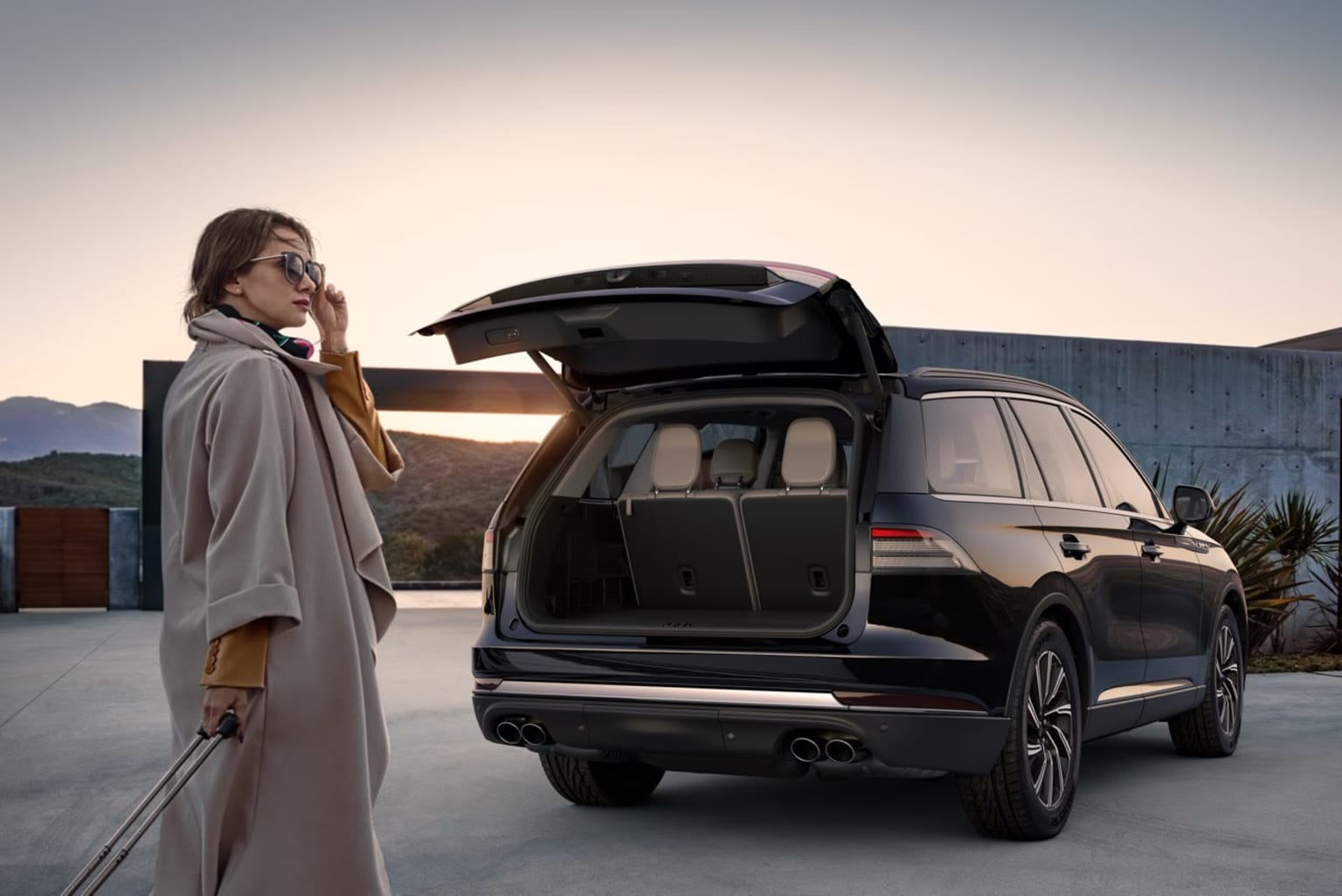 A person in a long coat stands next to a large SUV with its trunk open, against a backdrop of mountains and a clear sky.