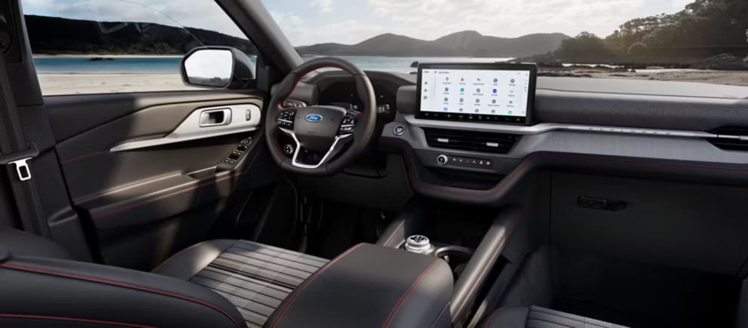 Interior of a modern vehicle with a large touchscreen display, steering wheel, and gear shift, set against a scenic backdrop of mountains and a body of water.