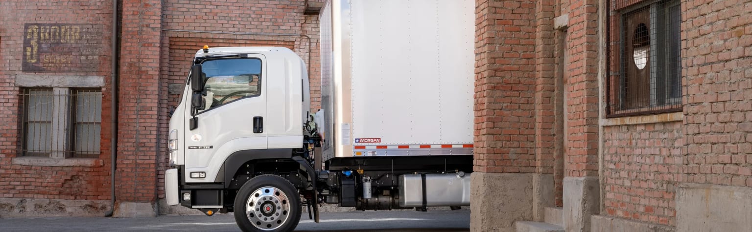 A white delivery truck parked in a narrow alley between two brick buildings, with the truck's rear cargo area visible.