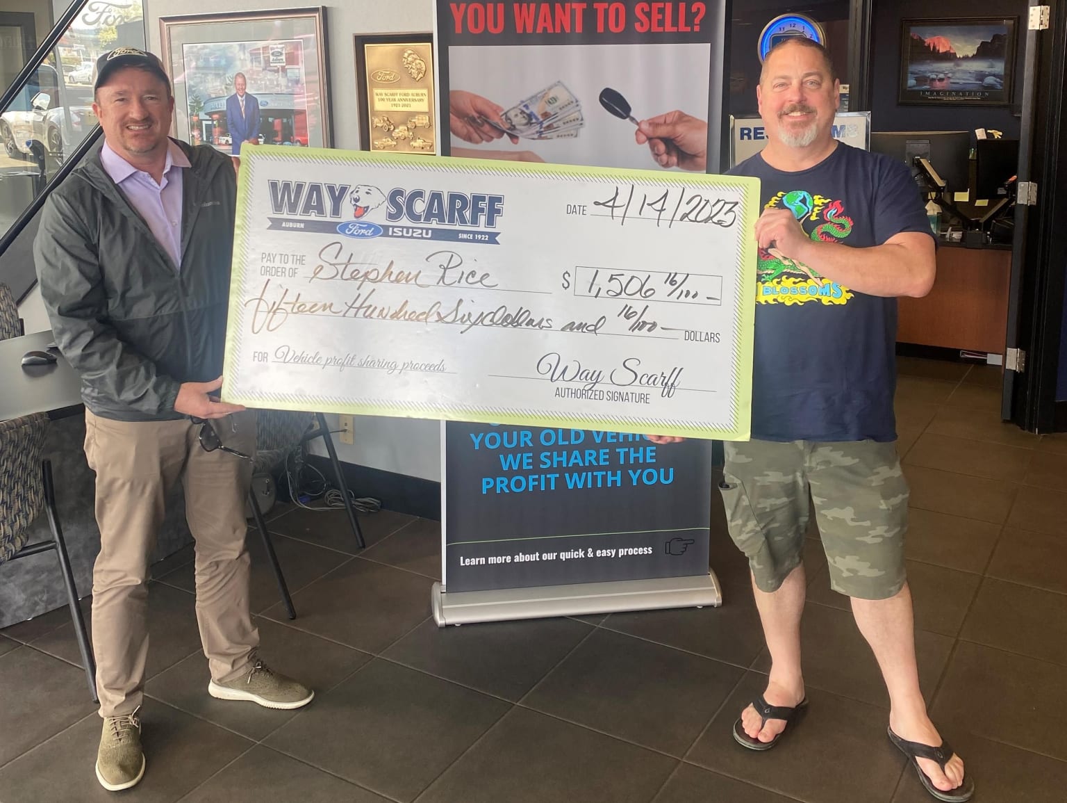 Two men standing in a business setting, one holding a large check and the other wearing a t-shirt and sandals, with a sign advertising "You want to sell?" and "We share the profit with you" in the background.