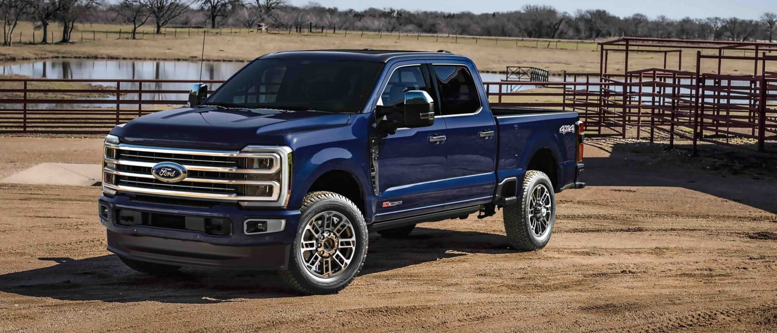 A dark blue truck from the F-Series is parked on a dirt lot with a fenced pasture and bare trees in the background.