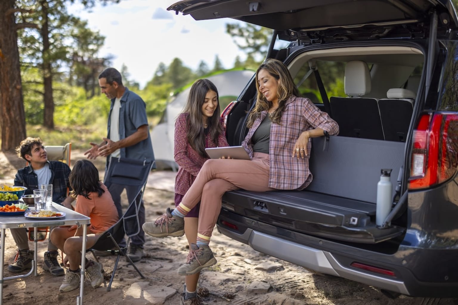 A group of people, including two women and a man, are gathered around a vehicle in a wooded outdoor setting, with a table set up nearby and a child visible in the background.