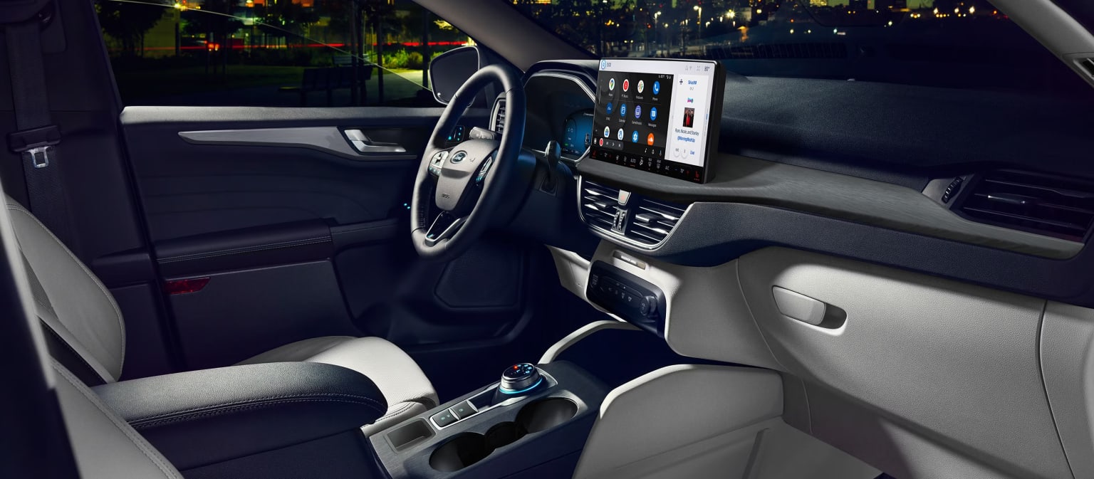 The image shows the interior of a modern car, with a steering wheel, dashboard, and various controls and displays visible. The background appears to be a nighttime urban setting, with the car's headlights illuminating the surroundings.