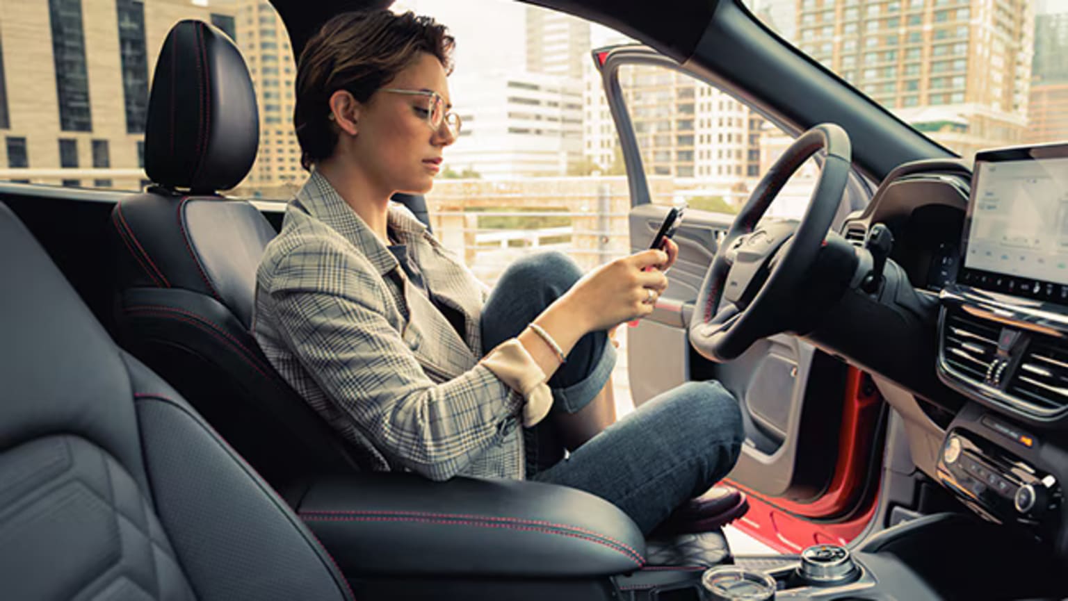 A person in a gray plaid jacket sitting in the driver's seat of a car with a cityscape visible through the windshield in the background.
