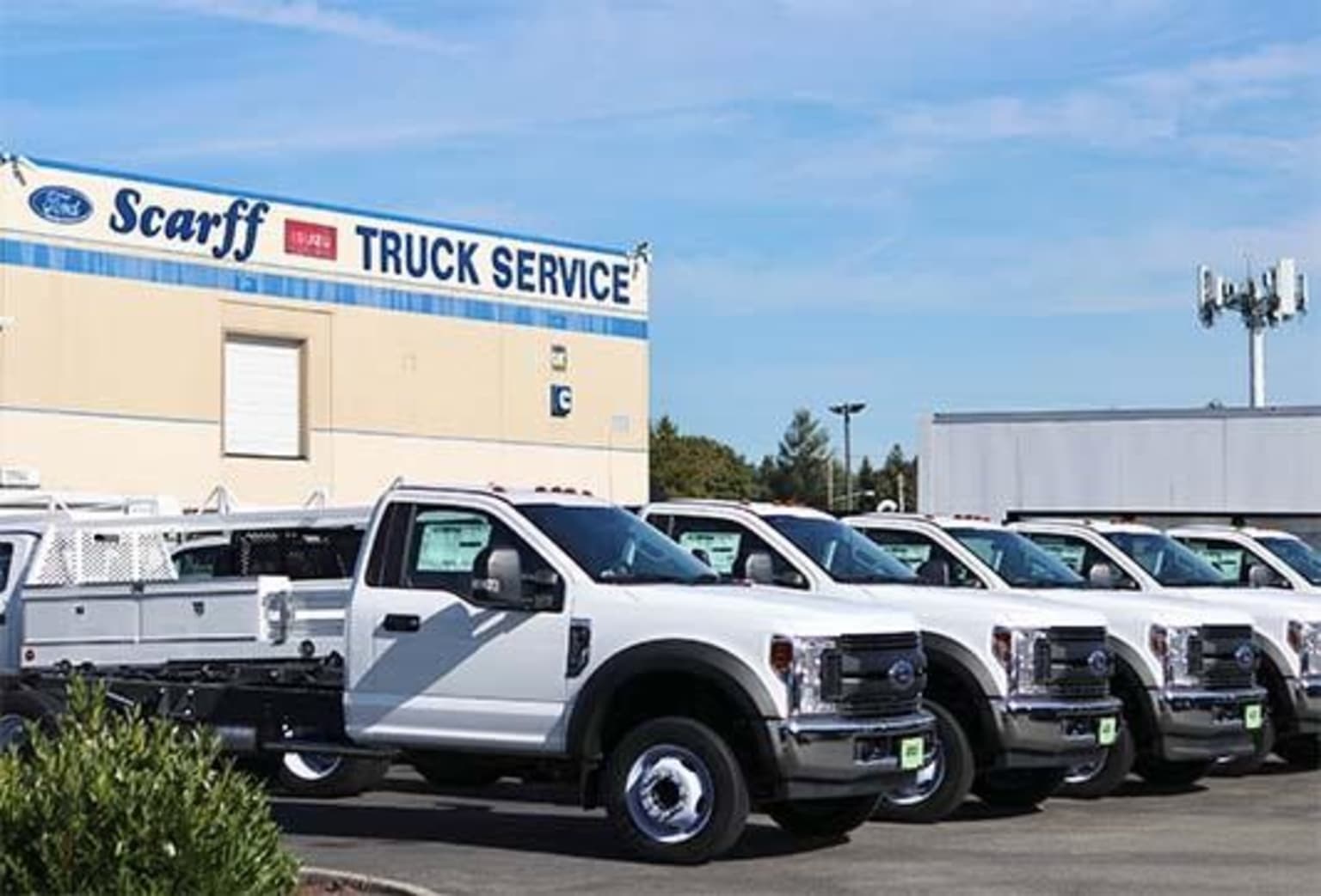 A row of white utility trucks parked in front of a truck service building, set against a blue sky with some trees in the foreground.