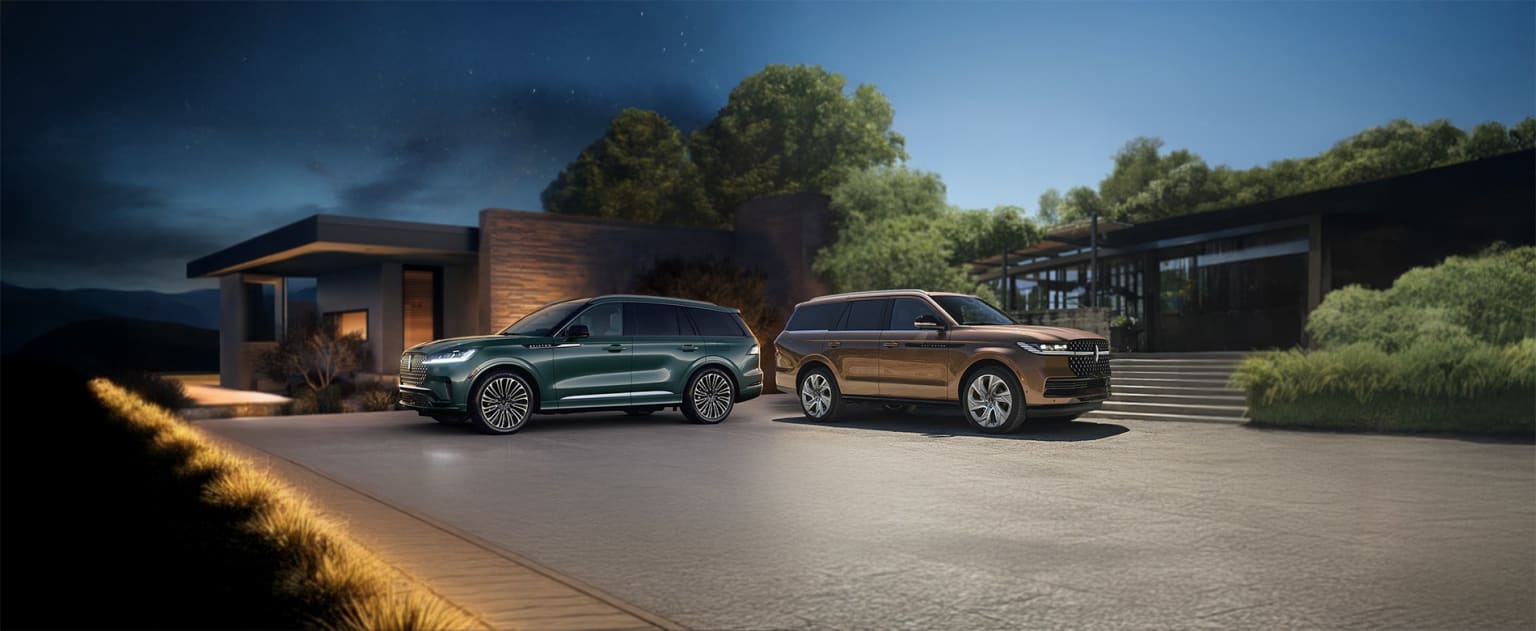 Two luxury SUVs parked in front of a modern building, surrounded by lush greenery and a clear blue sky. One SUV is green and the other is gold.