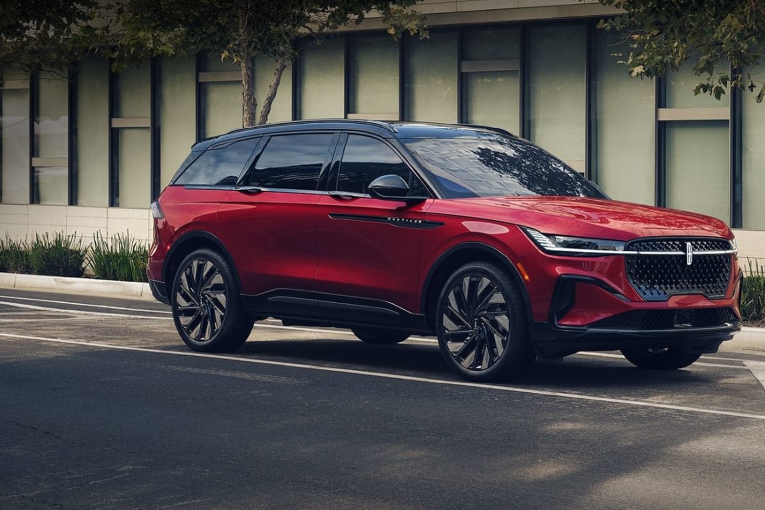 A sleek, red SUV with a bold, angular design parked on a paved street, surrounded by a modern architectural backdrop with large windows.