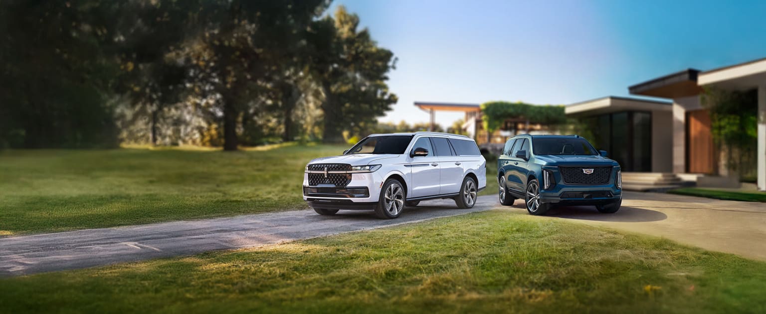 Luxury SUVs parked on a grassy driveway, surrounded by lush trees with a clear blue sky in the background.