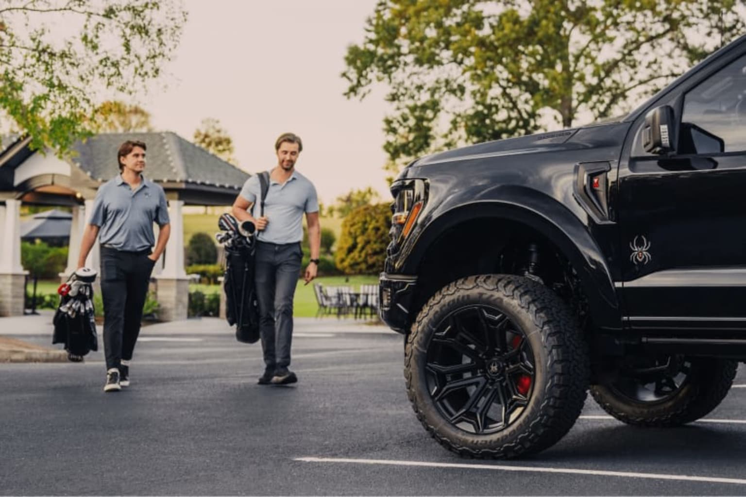 Two men carrying golf bags walking towards a black lifted pickup truck in a parking lot.