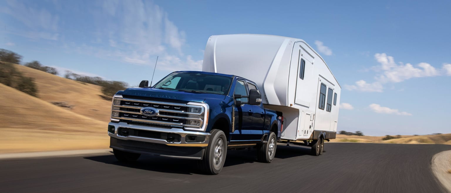 A large blue pickup truck is towing a white recreational vehicle on a dusty road against a backdrop of a clear blue sky.