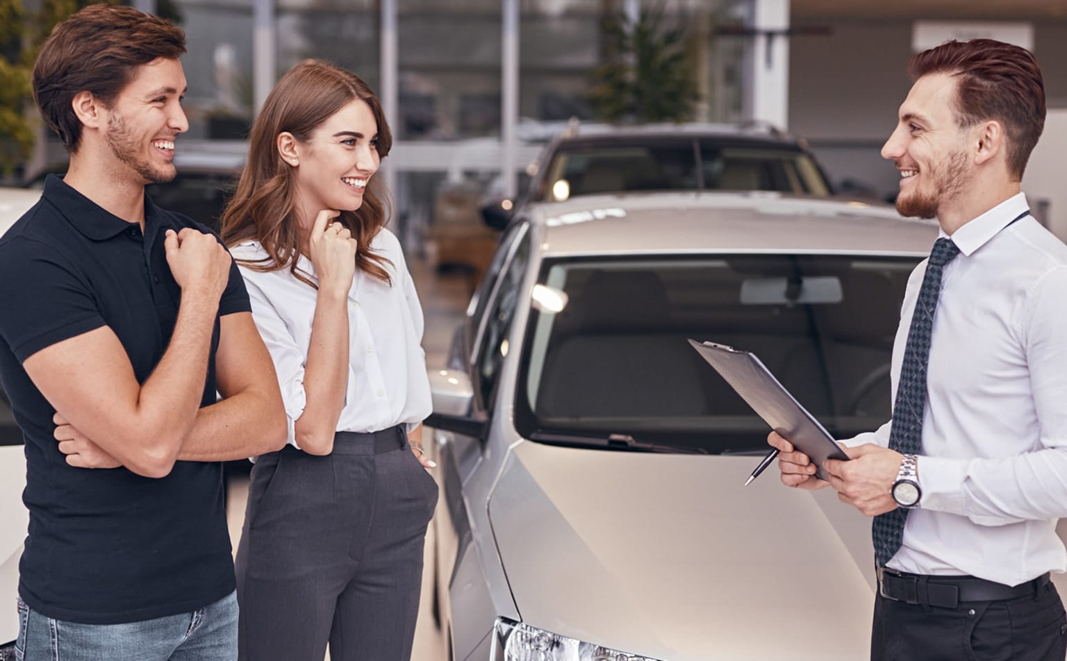 A smiling couple with a car salesman in a brightly lit showroom filled with cars.