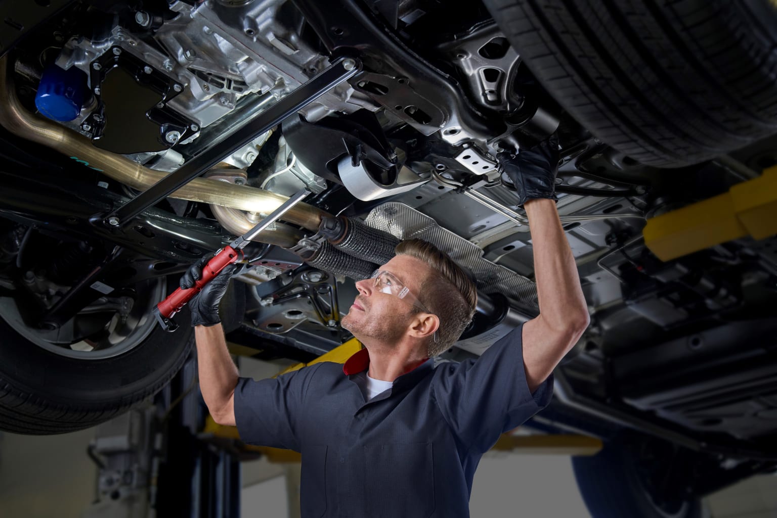 A mechanic working on a car's engine in a garage, holding a wrench and working on the car's hood.