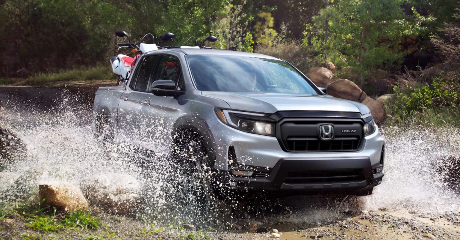A silver truck with a motorcycle in its bed splashes through a shallow stream with a wooded hillside in the background.