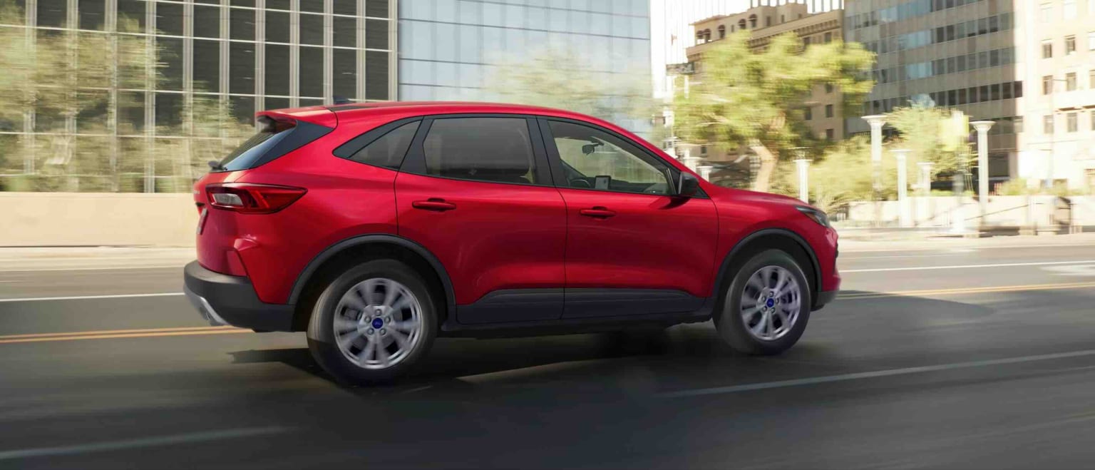 A red SUV drives down a city street with modern buildings in the background.