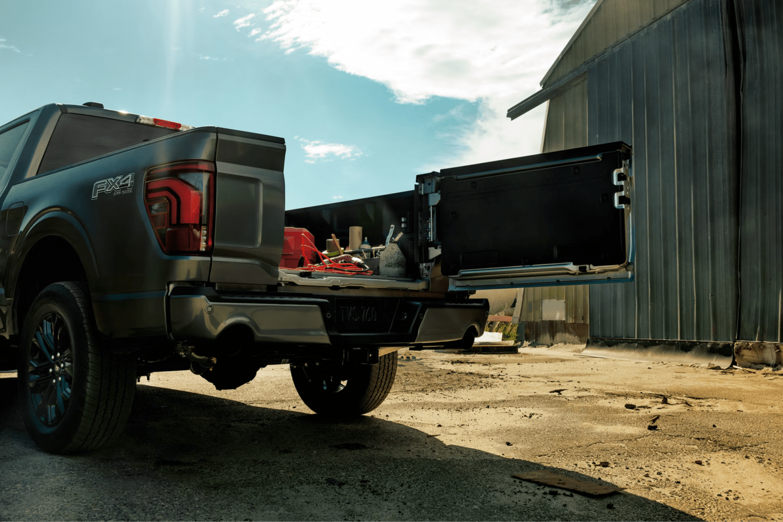 A pickup truck with an open storage drawer full of tools sits in the foreground against a rustic metal building and a bright, cloudy sky.