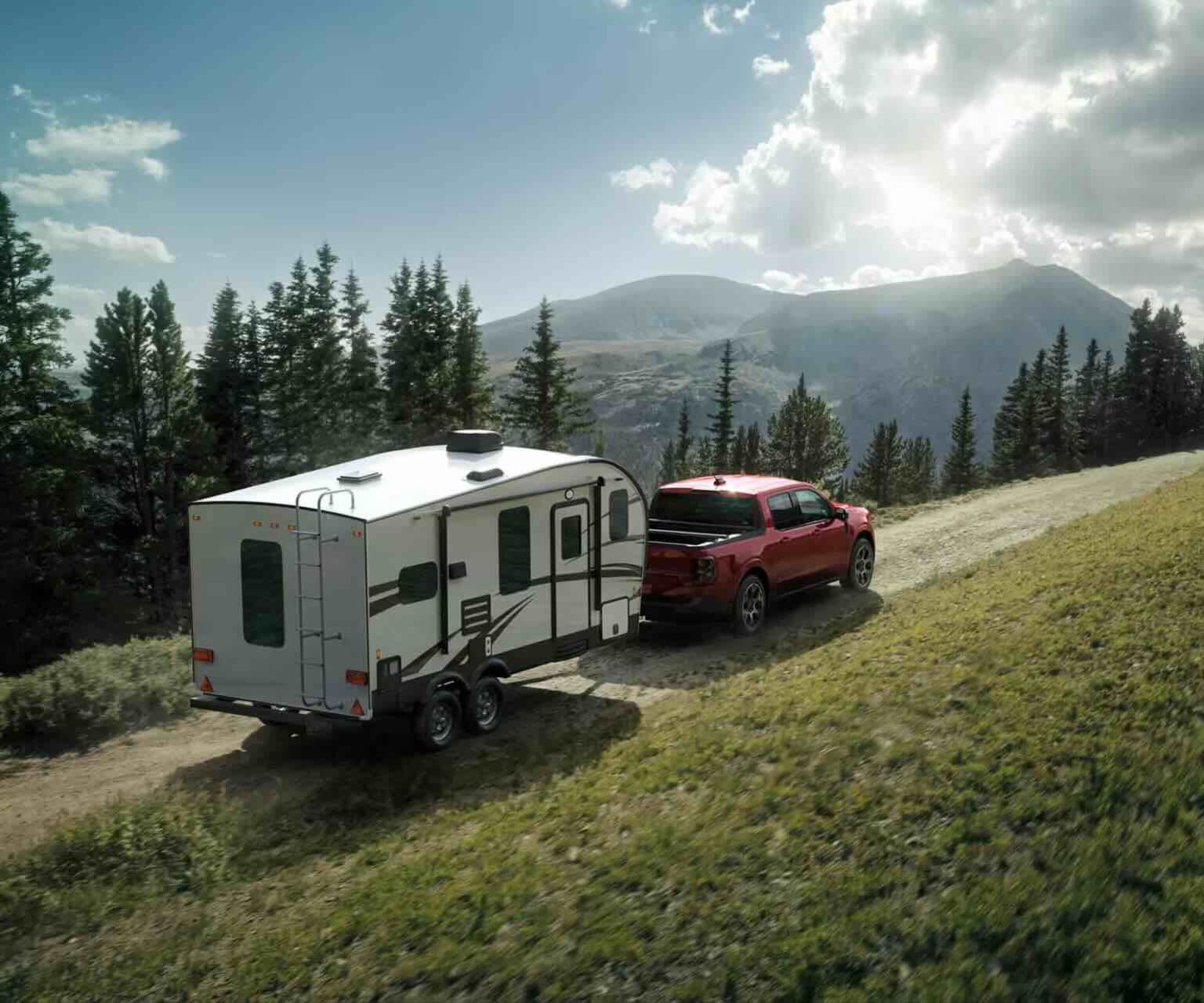 A red pickup truck towing a white travel trailer through a forest on a dirt road, with mountains in the distance under a bright sky.