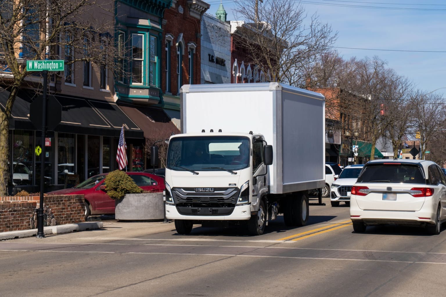 A white box truck drives down a street lined with brick buildings, with a red sports car and other vehicles visible in the background.