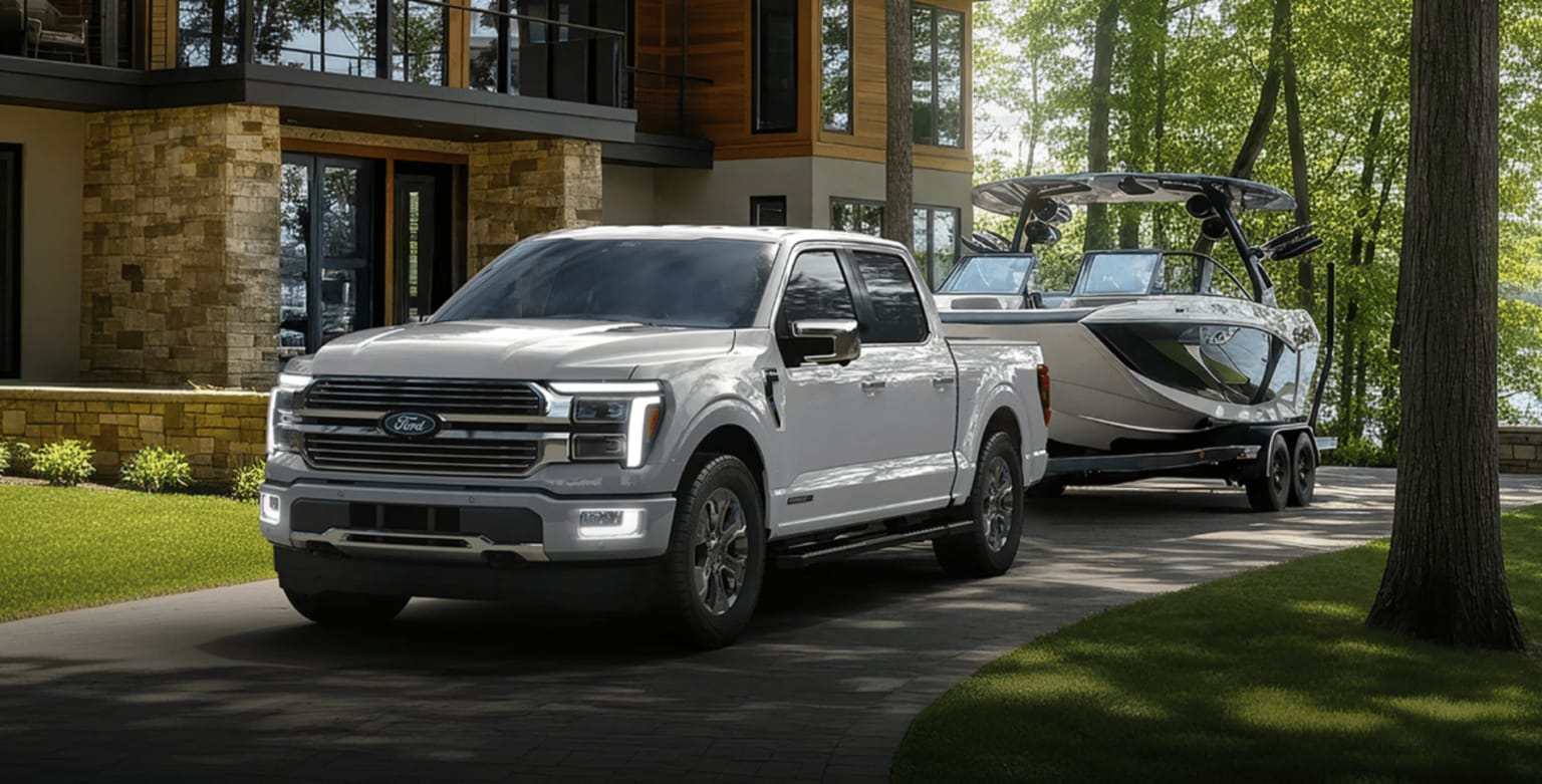 A white pickup truck towing a boat sits on a paved driveway in front of a modern stone house and lush green trees.