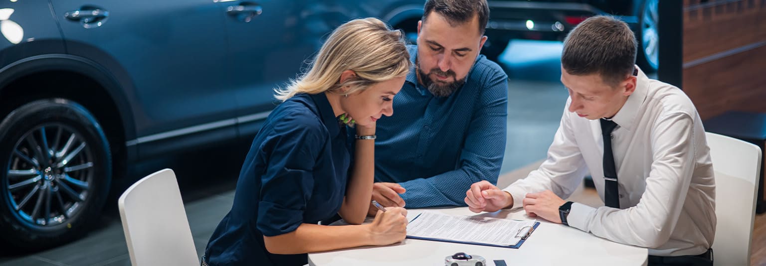 A group of people, including a man and a woman, are gathered around a table, engaged in what appears to be a professional or business discussion, with a computer monitor visible in the background.