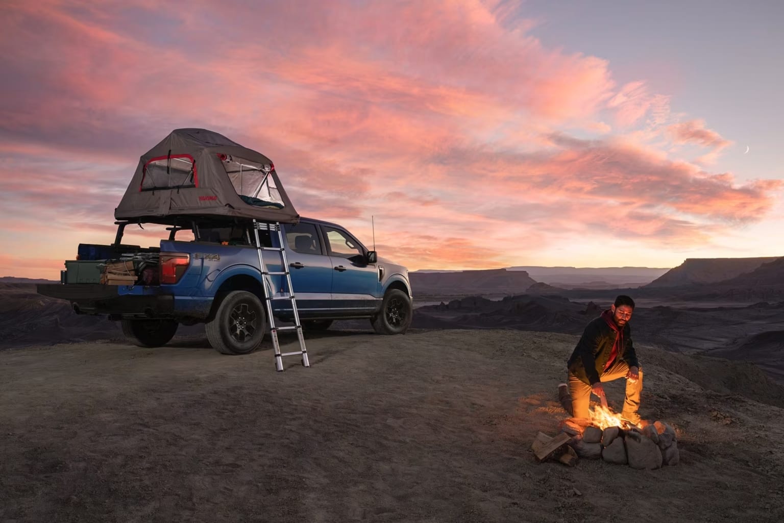 Off-road vehicle with rooftop tent in desert landscape at sunset with person by campfire.