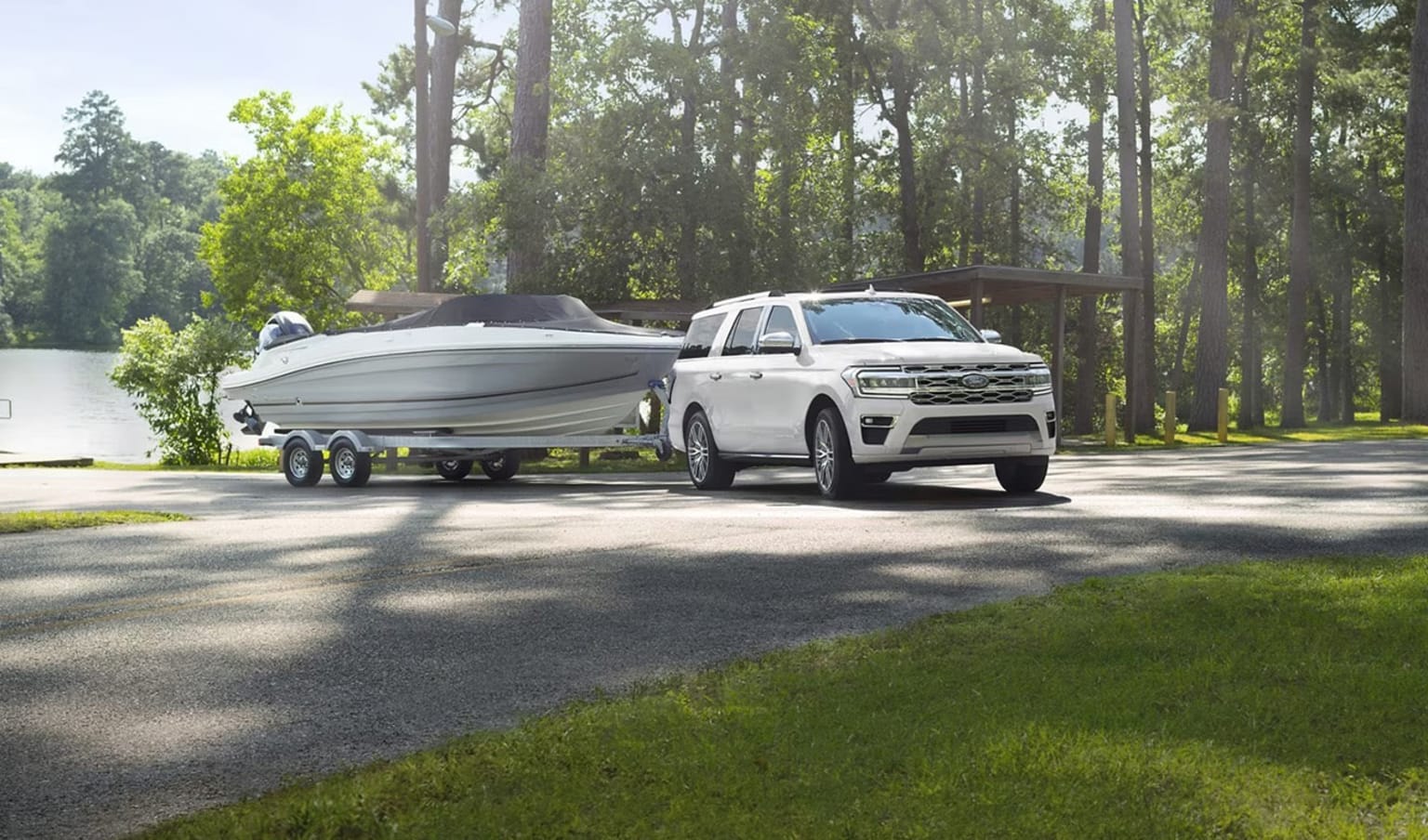 A large white SUV towing a boat on a trailer through a wooded area, with lush green trees and grass surrounding the scene.