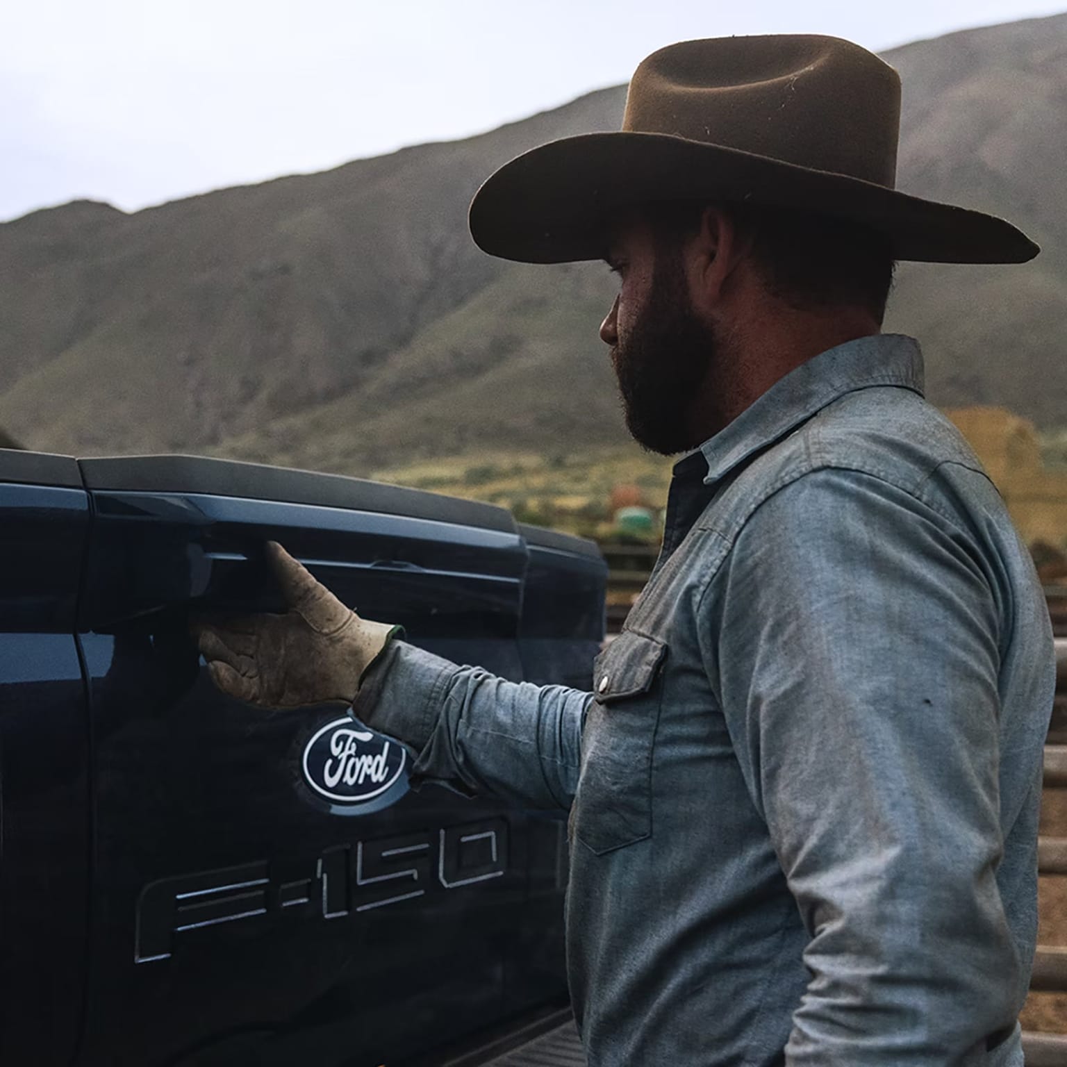 A man wearing a wide-brimmed hat and a denim jacket stands next to a pickup truck, with a mountainous landscape visible in the background.