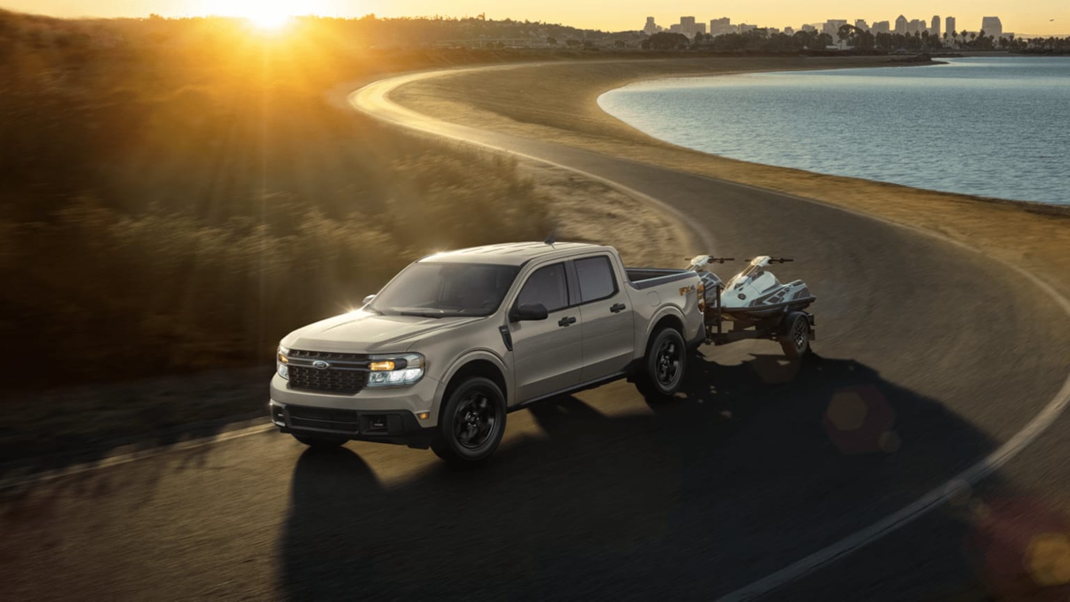 A silver pickup truck on a coastal road at sunset with a city skyline in the background.