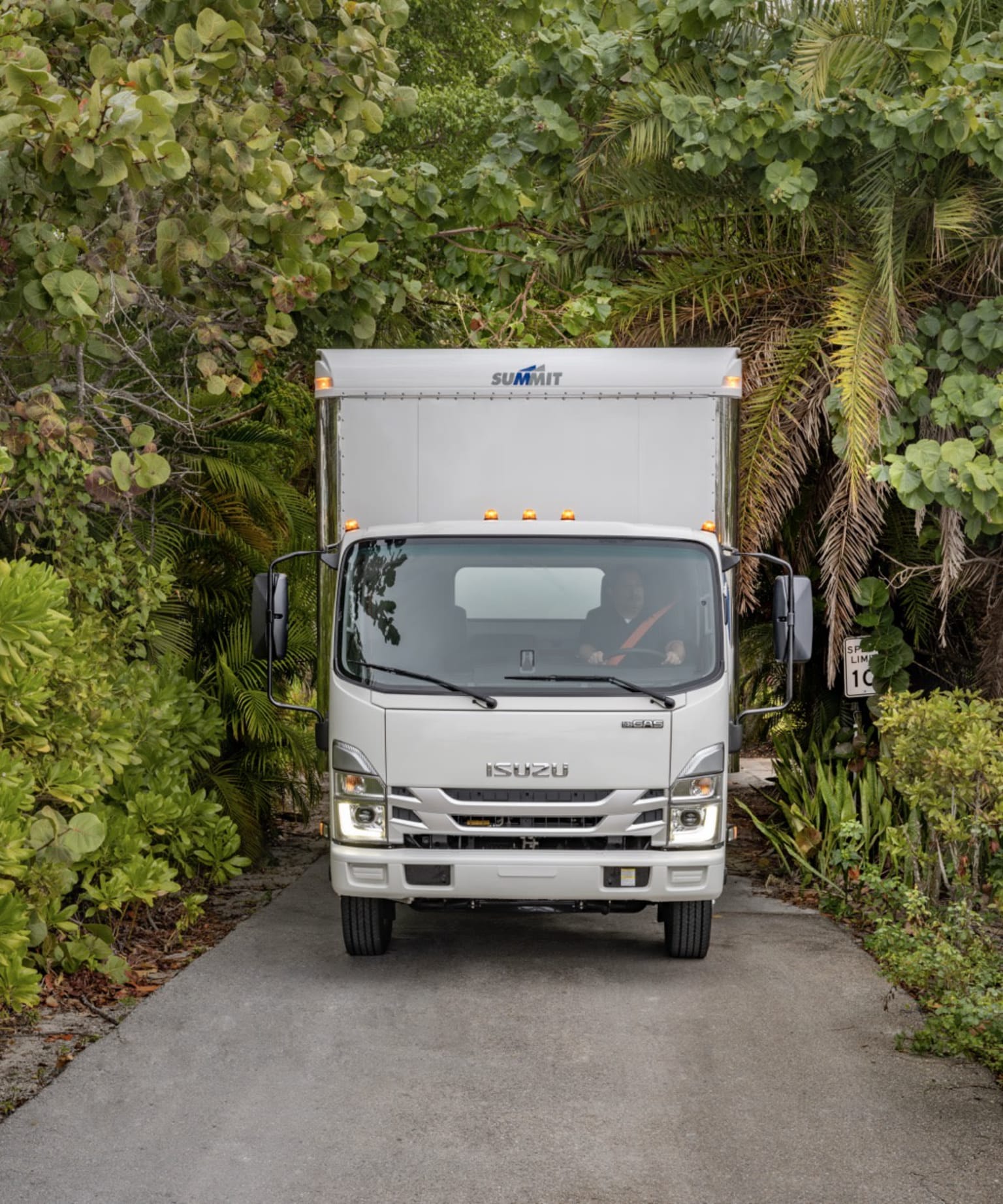A white delivery truck parked on a paved driveway surrounded by lush, green foliage and vegetation.