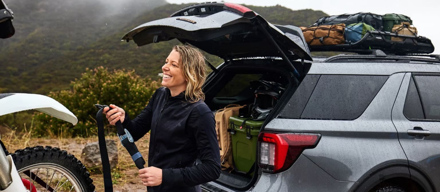 A young woman stands next to an open car trunk, surrounded by outdoor scenery with mountains and vegetation in the background.