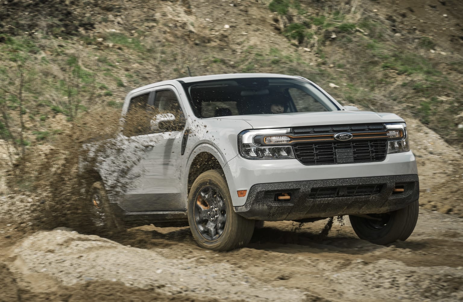 a white pickup truck driving through a mud covered hill in the mud on a dirt road with a hill in the background